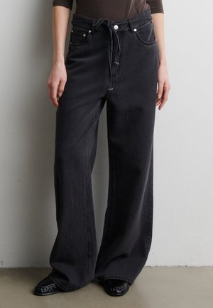 Personne portant un jean noir taille haute à jambes larges avec un cordon à la taille et des chaussures noires, debout contre un mur clair uni.
