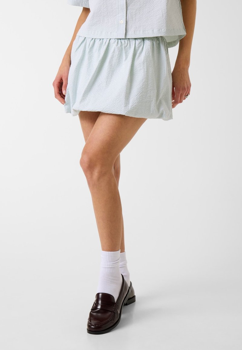 Femme portant une mini-jupe texturée bleu clair, un haut boutonné assorti, des chaussettes blanches et des mocassins en cuir brun foncé, debout devant un fond blanc.