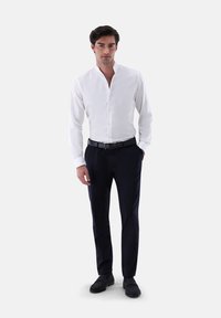 Weißes Langarmhemd mit Stehkragen, kombiniert mit dunklen Hosen. Das Modell trägt einen schwarzen Gürtel und dunkle Wildlederschuhe. Schlichtes, maßgeschneidertes Design.