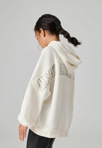 Hoodie oversize color crema con cappuccio regolabile e dettaglio testuale decorativo con borchie sul retro. Tessuto morbido e orlo a coste. Maniche lunghe.