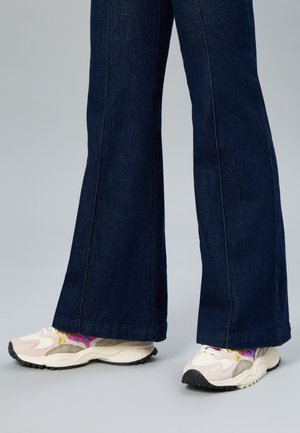 Person trägt ausgestellte dunkle Jeans aus Denim mit weißen und beigen klobigen Sneakern und bunten gemusterten Socken auf grauem Boden.