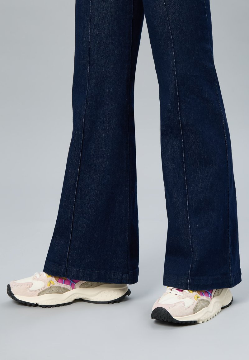 Personne portant un jean évasé en denim foncé avec des baskets épaisses blanches et beiges et des chaussettes colorées à motifs sur un sol gris.