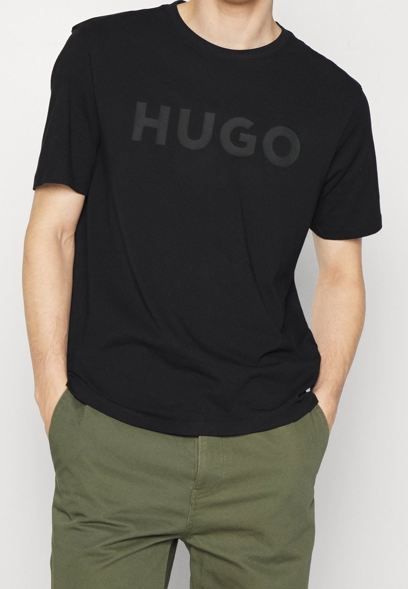 Man wearing black short-sleeve T-shirt with subtle "HUGO" text, paired with olive green pants, hands in pockets.