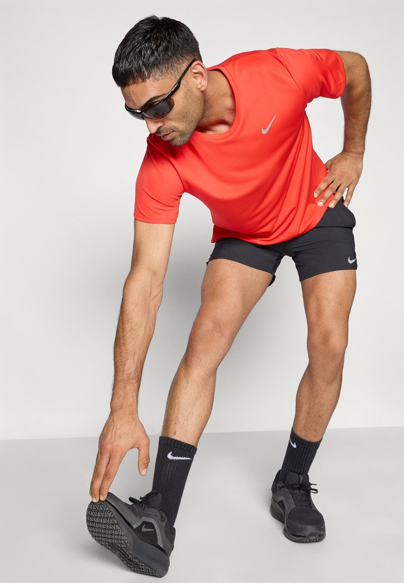Rotes sportliches T-Shirt, schwarze Shorts, schwarze Turnschuhe und schwarze Socken. Die Person dehnt sich und trägt eine Sonnenbrille. Nike-Logo sichtbar.