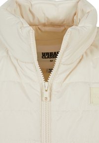 Veste matelassée beige clair avec un col montant, une fermeture éclair frontale et une texture matelassée horizontale. Elle comporte un patch logo sur le côté supérieur gauche.