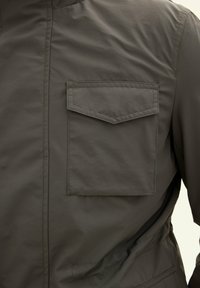 Giacca verde scuro con una leggera lucentezza, dotata di tasca sul petto, texture liscia e design minimalista. Dettagli cuciti visibili.