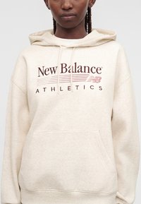 Personne avec des cheveux tressés portant un sweat à capuche beige clair New Balance Athletics avec une poche avant et une capuche à cordon.