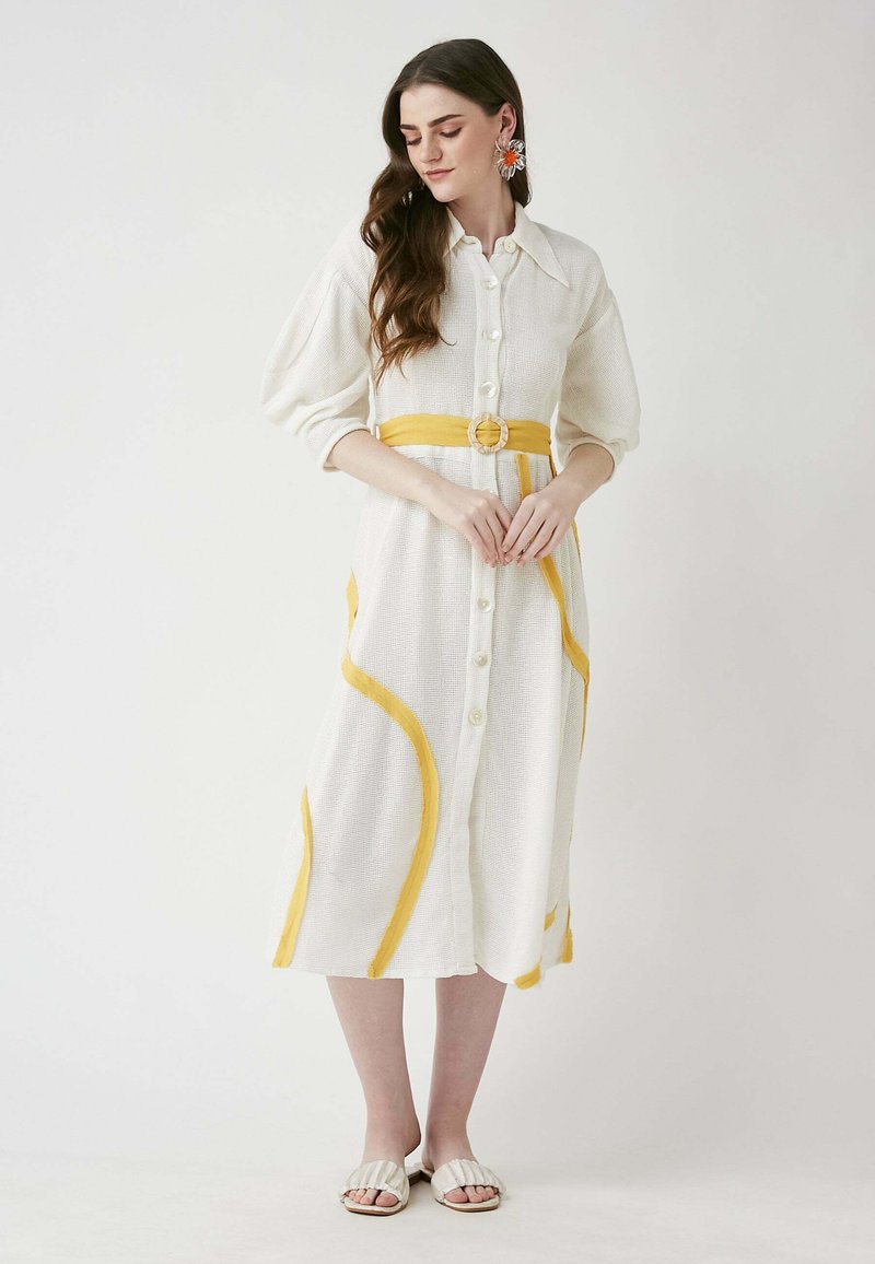 Weißes Kleid mit gelben Details, Knopfleiste und tailliertem Bund. Strukturiertes Gewebe, lange Ärmel und fließende Silhouette. Schlichter Hintergrund.