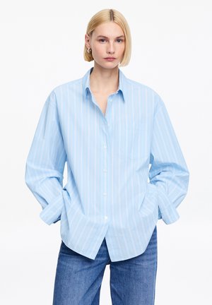 Mujer rubia con camisa de botones extragrande de rayas azul claro, con las manos en los bolsillos y vaqueros azules, sobre un fondo blanco.