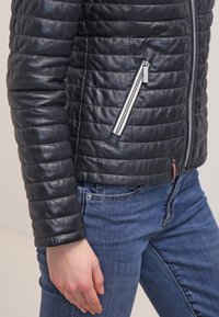 Zbliżenie osoby noszącej czarną pikowaną skórzaną kurtkę z ukośną kieszenią na zamek oraz niebieskie dżinsy.