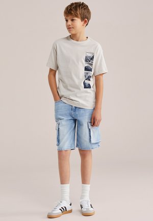 Fant deček stoji z rokami v žepih, oblečen v bež majico z grafičnim potiskom, svetlo modre kavbojske cargo kratke hlače, bele nogavice in bele superge s črnimi črtami.