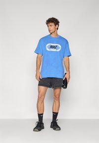 Blå bomulds T-shirt med et stiliseret sort og hvidt Nike-logo, parret med mørke shorts og sorte atletiske sko, som holder en sort vandflaske.