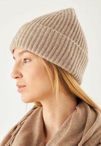 Chapeau en tricot côtelé beige avec une couronne pointue ; texture douce, éclat subtil. Accompagné d'une écharpe beige assortie en arrière-plan.