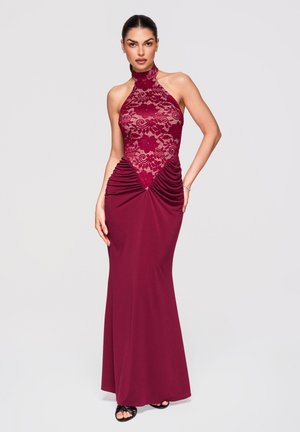 Femme portant une robe bordeaux sans manches, longue jusqu'au sol, avec un corsage en dentelle et des côtés froncés, debout devant un fond clair uni.