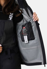 Sort vandtæt jakke med gråt indre, der har en lynlåslomme og mærkedetaljer på foret. Justerbare stropper og synligt metaludstyr.