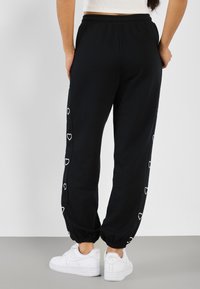 Kvinna i svarta joggers med vita hjärtan på sidorna och vita sneakers, stående med händerna bakom ryggen på en grå golv.