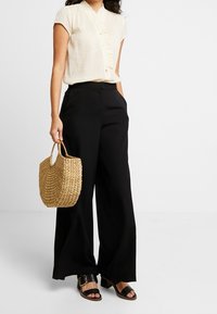 Blouse beige à manches courtes et boutonnée avec des rayures verticales, associée à un pantalon large noir. Tient un sac en paille tissé. Sandales noires.