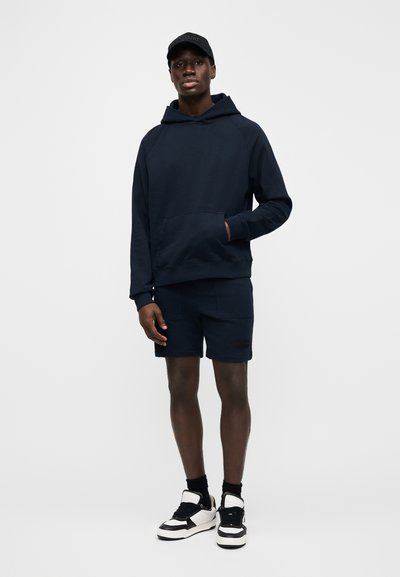 Námořnicky modrá bavlněná mikina s kapucí a přední kapsou, doplněná o sladěné námořnicky modré šortky. Černé a bílé tenisky. Jednoduchý design, bez výrazných vzorů.
