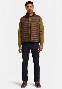 Bruine pufferbodywarmer met verticale stiksel, gedragen over een olijfgroene sweatshirt. Strak zittende donkere jeans en bruine verweerde schoenen maken de outfit compleet.