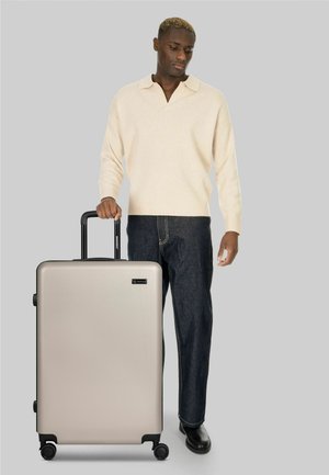 Uomo con maglione beige e pantaloni scuri in piedi che tiene il manico di una grande valigia rigida argento con quattro ruote.