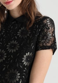 Femme portant un haut en dentelle noire avec des motifs floraux et un col noir uni, montrant un gros plan du haut du torse et d'une partie du visage.