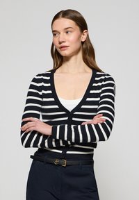 Randig kofta i svart och vitt med djup V-hals, ribbad muddar och knappstängning. Kombinerad med en vit topp och mörka byxor.