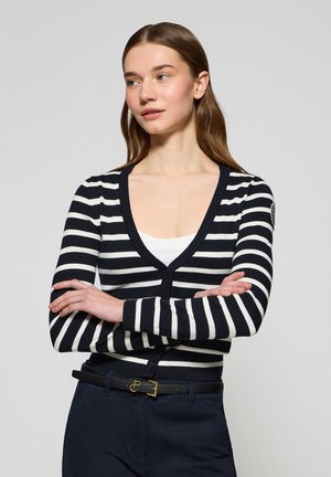 Striped cardigan in black and white with a deep V-neck, ribbed cuffs, and button closure. Paired with a white top and dark pants.