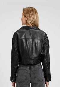 Schwarze Lederjacke in Crop-Form mit breitem Kragen, Reißverschlussärmeln und detailreicher Nahtführung entlang des Rückens. Getragen über grauen hochgeschnittenen Jeans.