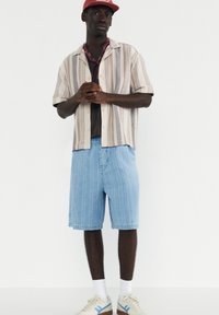 Chemise à manches courtes rayée en beige et marron, associée à un short en jean bleu clair. Les accessoires comprennent une casquette rouge et un foulard à motifs.