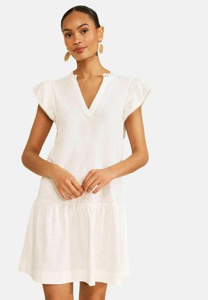 Kvinde iført en hvid kjole med korte ærmer, V-udskæring og flæset kant, kombineret med store runde vævede øreringe.