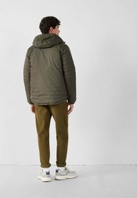 Uomo in piedi di fronte a una parete bianca, che indossa una giacca imbottita verde oliva, pantaloni marroni, calzettoni bianchi e sneaker bianche con dettagli grigi.