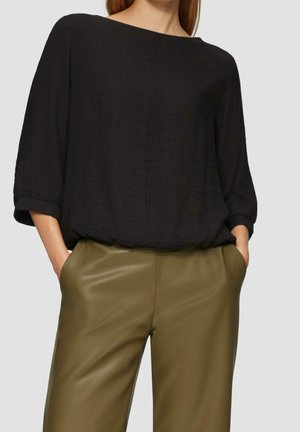 Femme portant un haut noir ample à manches longues rentré dans un pantalon en cuir vert olive taille haute, les mains dans les poches, debout devant un fond clair.