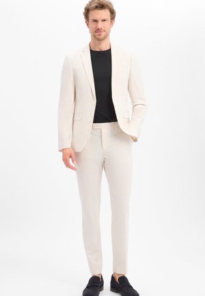 Mann trägt einen hellbeigen Anzug mit schwarzem Hemd und schwarzen Loafers, steht vor einem schlichten weißen Hintergrund.