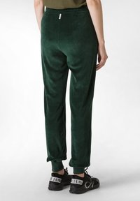 Grüne Samtjogginghosen mit elastischen Bündchen und einem Gummizug. Glatte Textur; minimales Design mit einem kleinen Etikett auf der Rückseite.