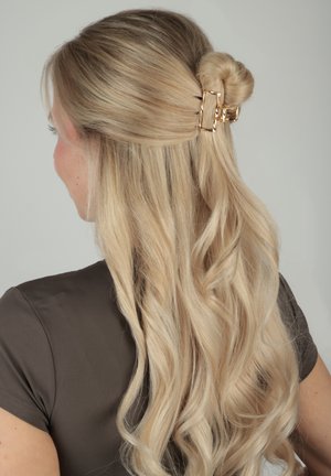 Donna bionda con capelli lunghi e ondulati, raccolti a metà e fissati con una clip rettangolare dorata, indossa una maglietta a maniche corte marrone scuro, vista di spalle.