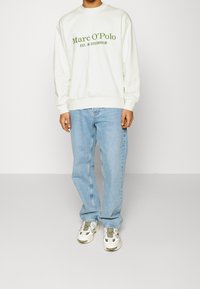 Sweat-shirt blanc avec texte vert "Marc O'Polo EST. À STOCKHOLM", associé à un jean bleu clair et des baskets blanches. Col rond, coupe décontractée.