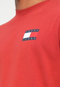 Červené bavlněné tričko s malým obdélníkovým logem Tommy Jeans v námořnické modři a bílé barvě na levé části hrudníku. Standardní kulatý výstřih.