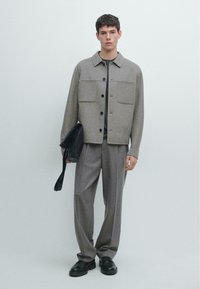 Lysegrå uldjakke med to forlommer, knaplukning og en rundet krave. Kombineret med grå plisserede bukser og sort fodtøj.