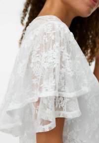 Haut en dentelle blanche doté de manches à volants superposées, de motifs floraux complexes et d'une finition légère et texturée.