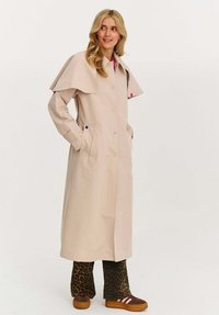 Žena v béžovém dlouhém plášti s capes detaily, leopardích kalhotách a hnědých teniskách, stojící s rukama v kapsách pláště.