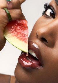 Zbliżenie na usta kobiety, błyszczące i ciemne, trzymające mokry plaster arbuza. Zielony pierścionek na dłoni. Kropla soku na ustach.