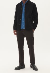 Black corduroy jacket with button closure over a blue denim shirt. Dark brown pants and black slip-on shoes complete the outfit.