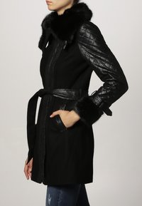 Donna che indossa un cappotto nero con maniche di pelle, colletto di pelliccia e polsini di pelliccia, con le mani in tasca, mostrata di lato su uno sfondo semplice.
