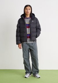 Black puffer jacket with hood, featuring a white logo. Worn over a gray striped sweatshirt and gray loose pants, with white sneakers.