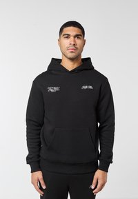 Muž v černé mikině s kapucí s bílým nápisem na hrudi, stojící před jednoduchým světle šedým pozadím, obrácený čelem k přední straně.