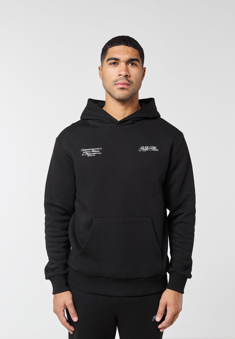 Muž v černé mikině s kapucí s bílým nápisem na hrudi, stojící před jednoduchým světle šedým pozadím, obrácený čelem k přední straně.