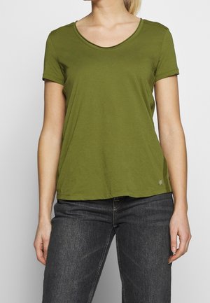 Vrouw draagt een effen olijfgroen shirt met korte mouwen en donkergrijze spijkerbroek, staand tegen een effen lichte achtergrond.