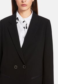 Black blazer with a structured design, featuring shiny buttons, subtle pocket details, and a white shirt with a graphic print underneath.