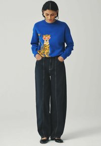 Niebieski sweter dziergany z graficznym motywem tygrysa z przodu, połączony z czarnymi dżinsami w stylu wide-leg z wysokim stanem, wyróżniającymi się widocznymi szwami.