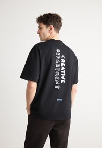 Black cotton t-shirt with short sleeves featuring vertical white text reading "CREATIVE DEPARTMENT" and a blue logo on the lower back.